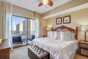 A hotel room with a large bed, a wooden headboard, a bench, a nightstand, and a balcony with a view of tall buildings.
