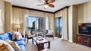 Habitación de hotel con sofá azul, una mesita de centro, televisor y balcón con vistas a la ciudad.