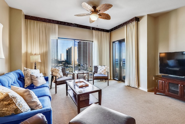 A hotel room with a blue sofa, a coffee table, a TV, and a balcony with a city view.