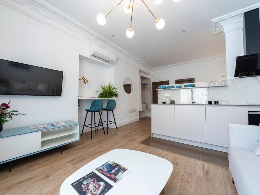 Una sala de estar moderna con televisor de pantalla plana, un sofá blanco, una mesa de centro con revistas y una zona de cocina con gabinetes blancos y una campana extractora negra.