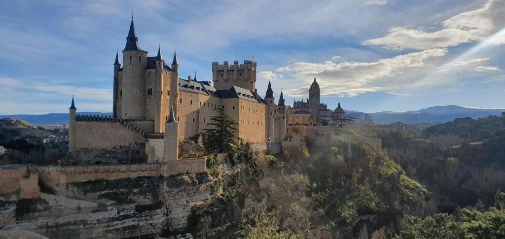 Un castillo histórico con múltiples torres y una muralla fortificada, rodeado de un paisaje montañoso.