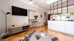 Un salon moderne doté d’un téléviseur à écran plat, d’un coin repas agrémenté d’un tapis à motifs zébrés et d’une cuisine avec des meubles blancs et une fenêtre à cadre noir.