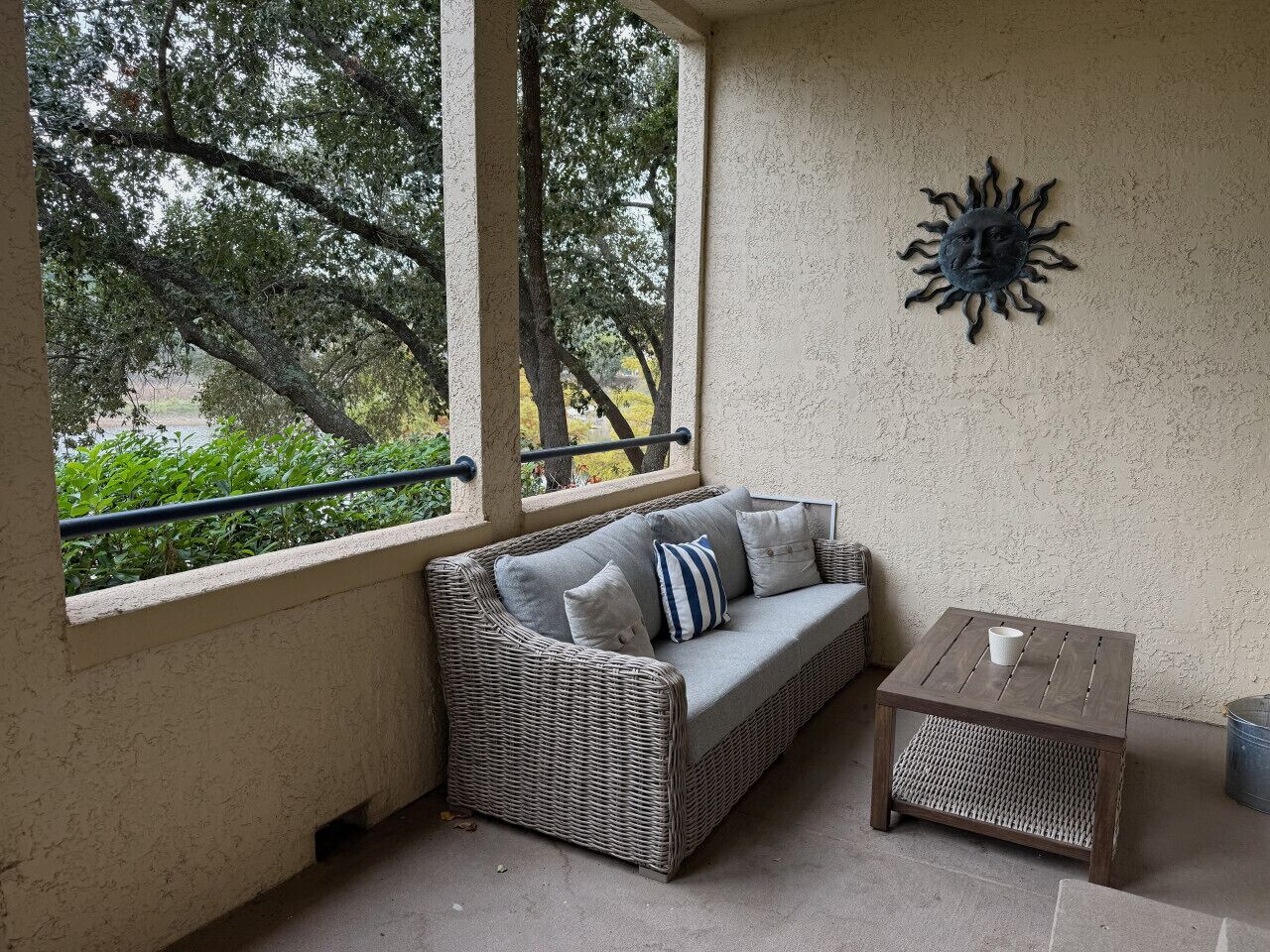A wicker sofa with cushions, a small wooden table, and a decorative wall piece in a covered outdoor area with a view of trees.