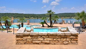 A pool area with a stone wall, palm trees, and a view of a body of water. - `Lakeside Haven`- Hot Tub, Pool, Lake view! (Lago Vista)