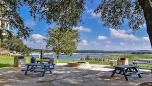 An outdoor patio with stone flooring, picnic tables, a fire pit, and a view of a lake and distant hills. - `Lakeside Haven`- Hot Tub, Pool, Lake view! (Lago Vista)