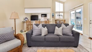 A living room with a grey sofa, a wicker chair, a lamp, and a dining area with a TV and chairs.