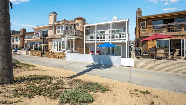 A beachfront property with a modern two-story house, a covered patio with outdoor seating, and a sandy beach in the foreground.