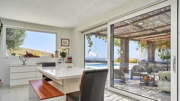 A modern kitchen with a large window, a dining area with a white table and chairs, and a view of an outdoor pool area. - Villa Spectacular Casa Negra with Sea View, Pool, Garden & Wi-Fi (Torrox Costa)