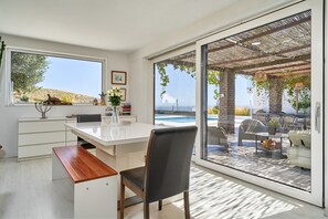 Une cuisine moderne dotée d’une grande fenêtre, un coin repas avec une table et des chaises blanches, et une vue sur une piscine extérieure.