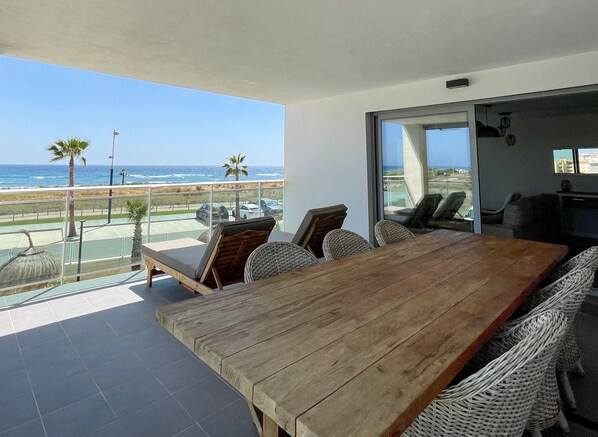 A wooden dining table with wicker chairs, a balcony with a view of the ocean, and a glass door leading to an indoor area.