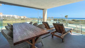 Un balcón con una mesa de madera y sillas de mimbre, con vistas a una zona costera con edificios y cielo despejado.