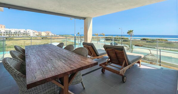 A balcony with a wooden table and wicker chairs, overlooking a coastal area with buildings and a clear sky.