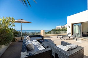 Una terraza en la azotea con mobiliario exterior, vista al mar y un edificio moderno al fondo.