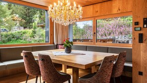 A dining area with a wooden table, patterned chairs, a chandelier, and large windows with a view of greenery.