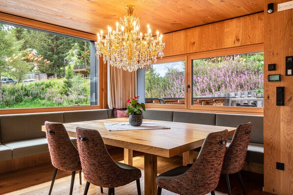 Un comedor con una mesa de madera, sillas estampadas, una araña y ventanas amplias con vistas a zonas verdes.