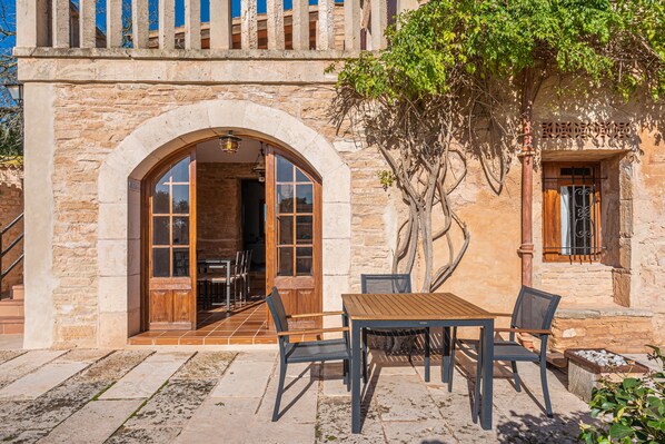 A stone building with an arched doorway, a wooden table with chairs, and a tree in front.