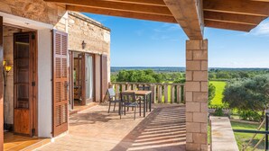 A covered patio with a table and chairs, a stone wall, and a view of a green landscape. - Apartment 'Es turo de Formentera' with Pool, A/C and Wi-Fi (Ses Salines)
