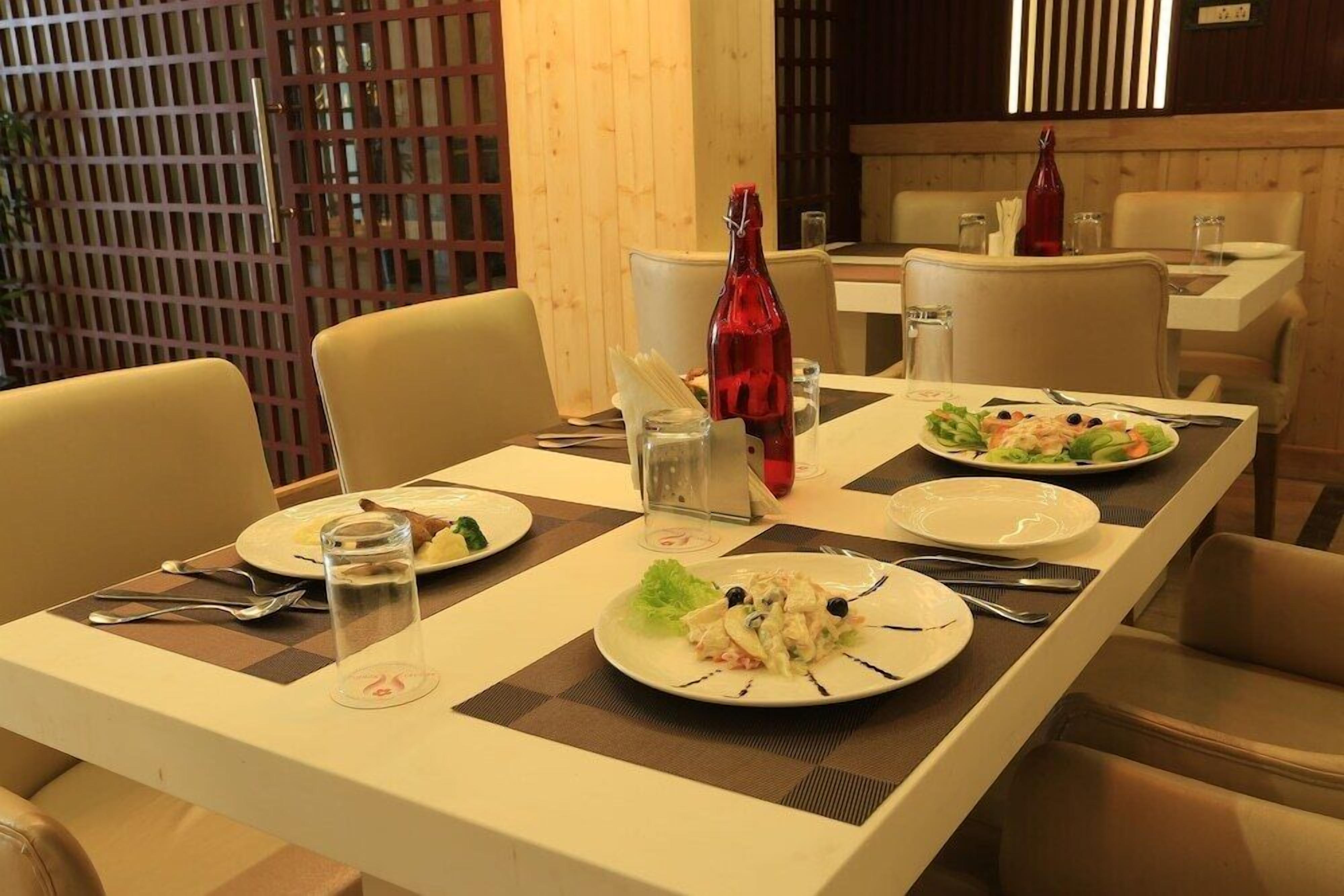Un coin repas avec une longue table dressée pour quatre personnes, comprenant des assiettes remplies de nourriture, des couverts et une bouteille de vin rouge.