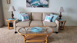 A living room with a beige sofa, two side tables with lamps, a glass coffee table, and a large seascape painting on the wall. - WOW! AMAZING OCEANFRONT CONDO 2nd Floor STUNNING VIEWS, GREAT LOCATION IN KAUAI (Kapaʻa)