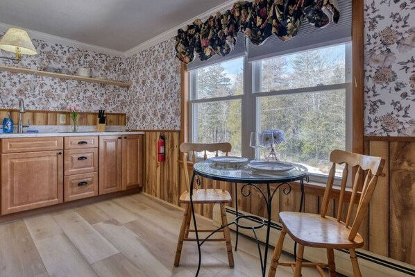 Una cocina con armarios de madera, una mesa redonda de vidrio y sillas. Hay una ventana con vistas a árboles y un extintor en la pared.