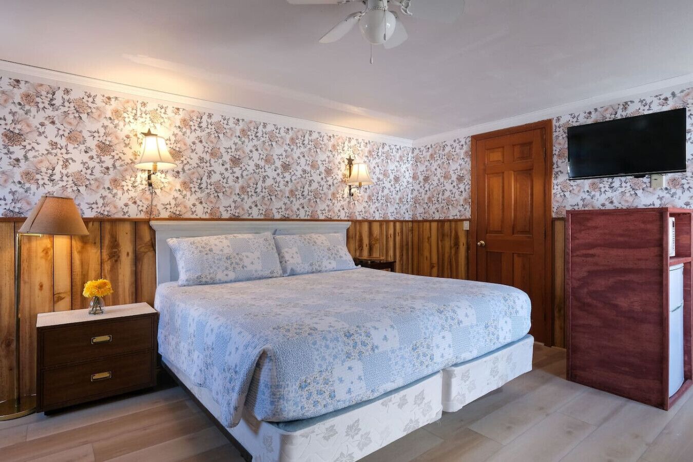 A bedroom with a bed, wooden headboard, two wall-mounted lamps, a TV, and a wooden wardrobe.