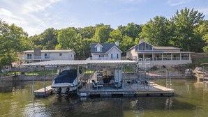A docked boat with a covered seating area, a small boat house, and a house with a balcony in the background. - Valley View 2 - Perfect for your next Lake Getaway! (Osage Beach)