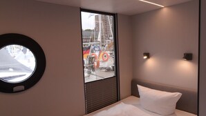 Quarto de hotel moderno com uma cama bem arrumada, uma janela redonda com vista para a cidade e uma luminária fixada na parede.