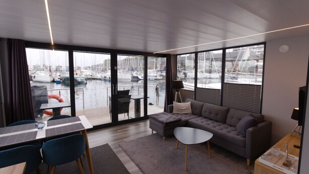 Uma sala de estar moderna com uma grande janela com vista para uma marina, um sofá cinzento, uma mesa redonda pequena e uma área de jantar com mesa e cadeiras.