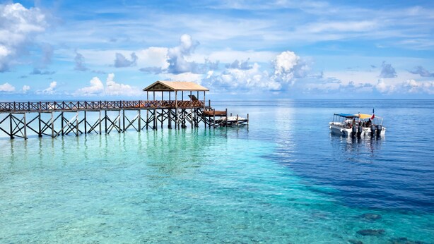 Eine hölzerne Pier erstreckt sich in klares türkisblaues Wasser, mit einem überdachten Sitzbereich und einem in der Nähe verankerten Boot.