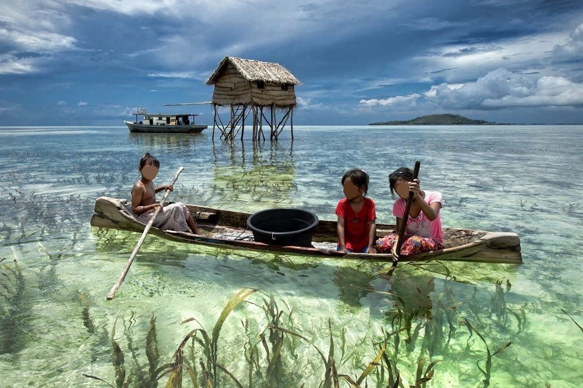 A clear tropical sea with a wooden boat carrying four people, a thatched hut on stilts, and a distant island.