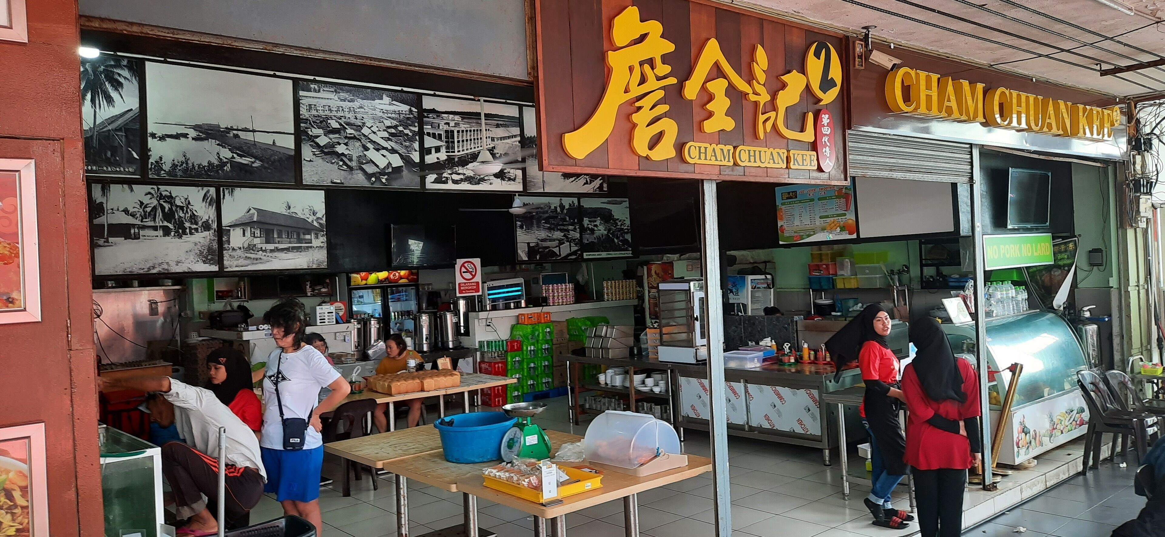 A restaurant with a sign reading "Cham Chuan Kee" and a menu board. There are tables and chairs inside, and a counter with a person serving food.