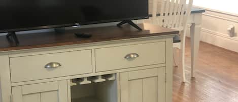 Uma sala de estar com uma televisão sobre um móvel de madeira, uma cadeira branca e um tapete estampado.