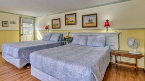 Una habitación con dos camas, una mesita de noche de madera con una lámpara, un ventilador y cuadros en la pared.