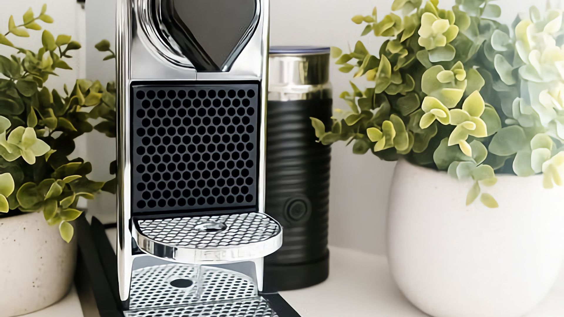 A coffee machine with a drip tray, a cylindrical container, and two potted plants.
