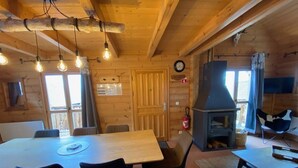En stuga med träpanel på insidan, med ett matbord, stolar, en öppen spis och en klocka på väggen.