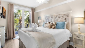 Habitación de hotel con una cama grande, un ventilador de techo, una mesita de noche, una lámpara y vistas a palmeras a través de la puerta corredera de cristal.