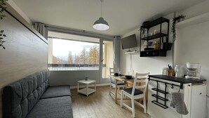 Un espace de vie compact comprenant un canapé, une table à manger, des chaises, un petit coin cuisine avec un réfrigérateur et un téléviseur fixé au-dessus du comptoir.