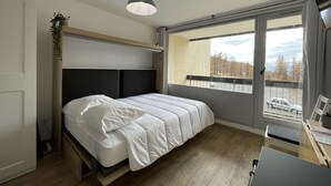 Une chambre avec un lit, un bureau, une lampe suspendue et une vue sur des arbres et un parking.