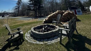 Een ronde vuurkuil met houtblokken, omringd door Adirondack-stoelen, gelegen in een grasrijk gebied met bomen op de achtergrond.