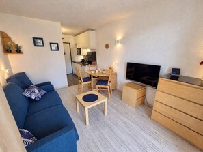 A compact living space with a sofa, a dining table, chairs, a television, and a small kitchen area.