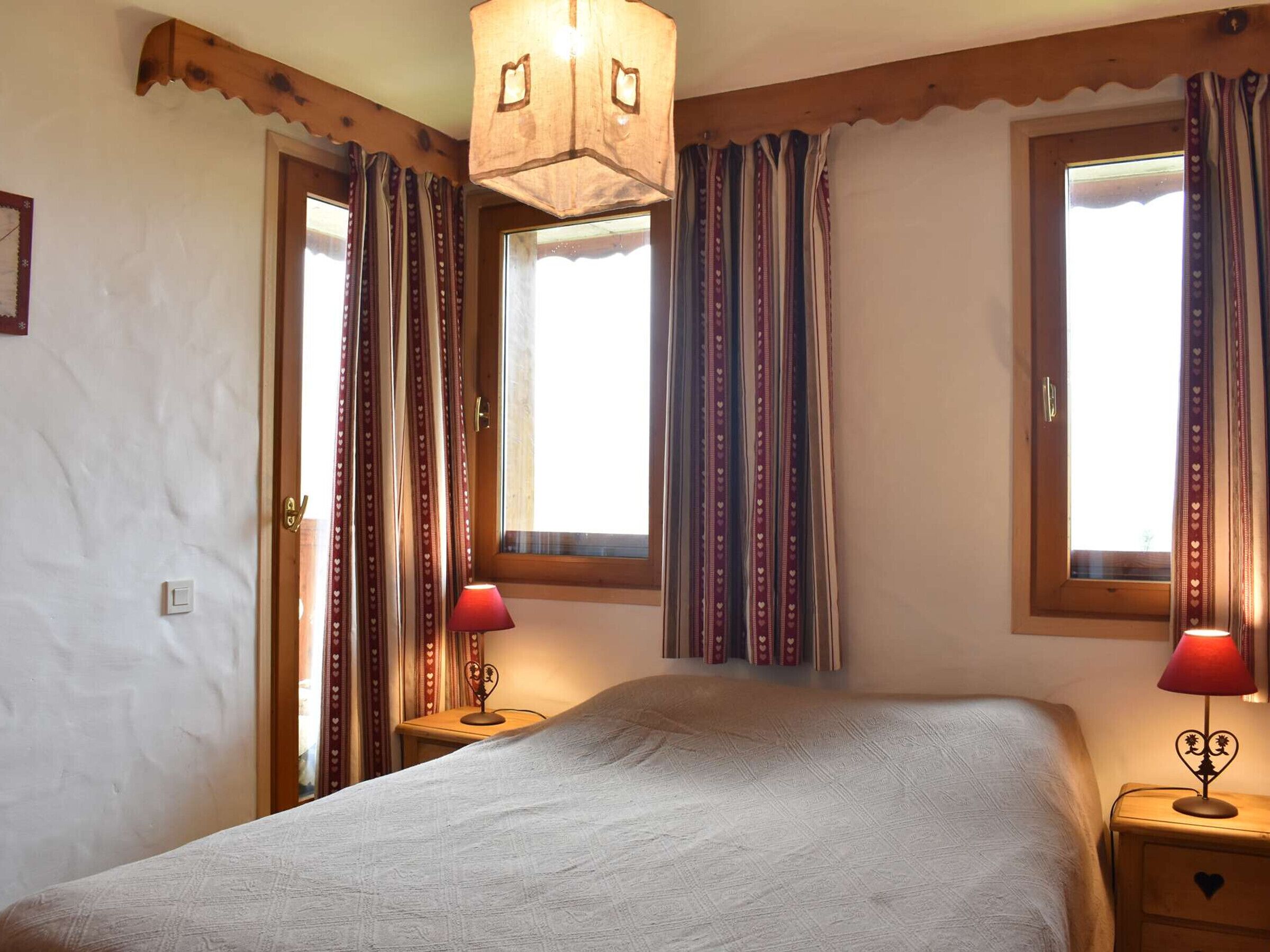 A bedroom with a bed, two bedside tables with lamps, a window with curtains, and a wooden ceiling light fixture.