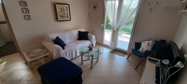 A living room with a white sofa, a glass coffee table, a blue ottoman, and a blue armchair. There is a framed picture on the wall, a window with curtains, and a door leading outside.