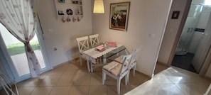 A dining area with a glass table, white chairs, and a lace tablecloth. There are wall-mounted shelves with plates and a framed picture.