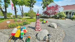 Un jardín con un faro blanco y rojo a rayas, boyas de colores y un edificio blanco con techo rojo.