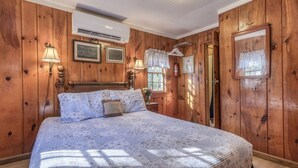 A wooden-paneled bedroom with a bed, framed pictures, and a window with lace curtains.