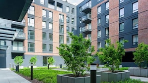A modern apartment building with multiple balconies, a landscaped courtyard with small trees, and a paved walkway.
