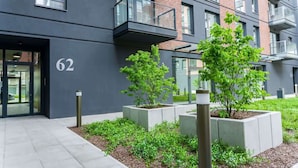 A modern building with a flat facade, numbered 62, featuring rectangular planters with small trees and shrubs, and a clean, paved entrance area.
