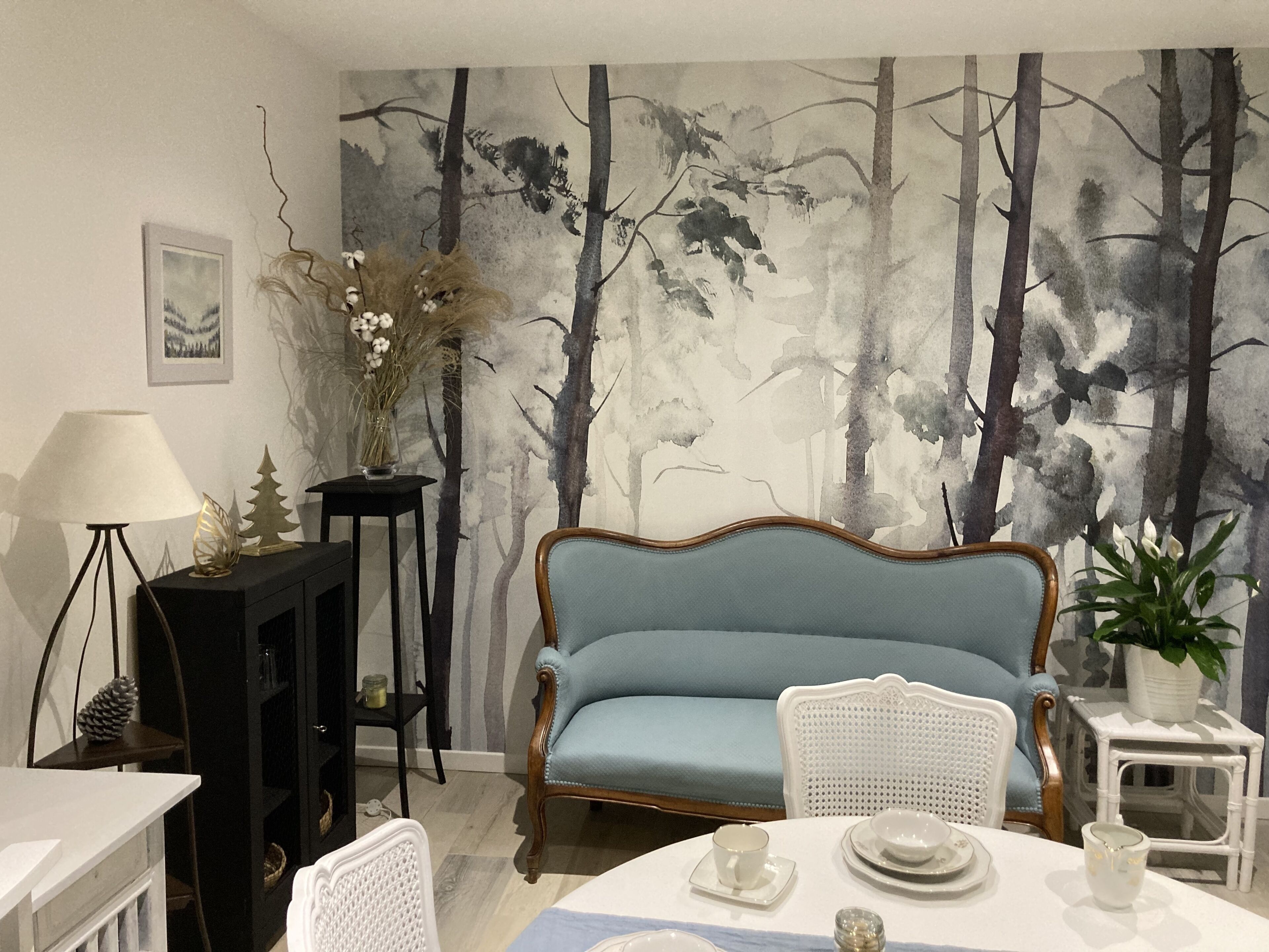 A dining area with a table set for two, a sofa, a lamp, and a wall mural.