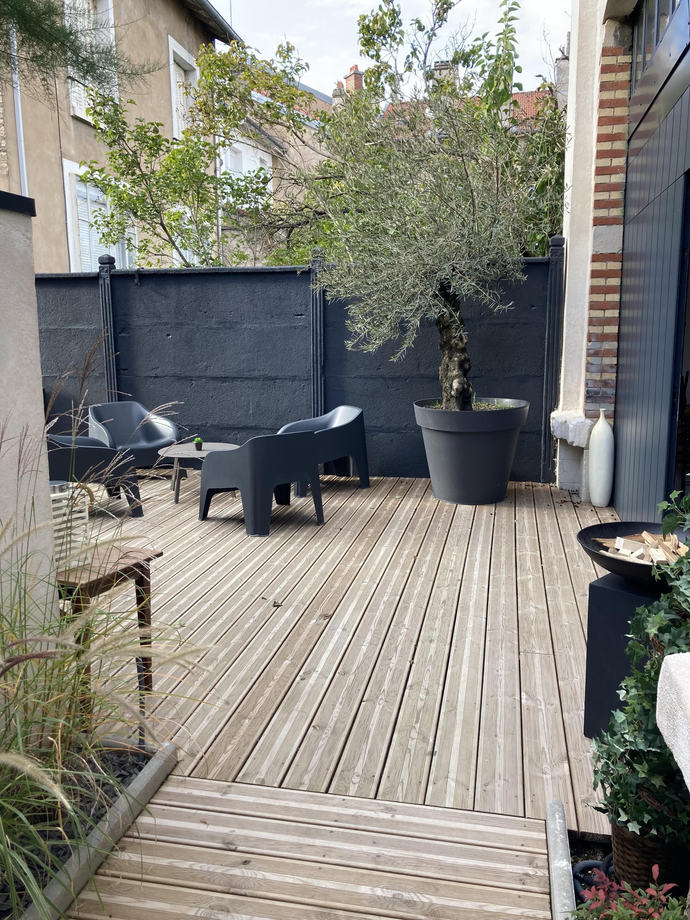 Um deck de madeira com móveis de exterior, uma grande árvore em vaso e uma parede de tijolos.