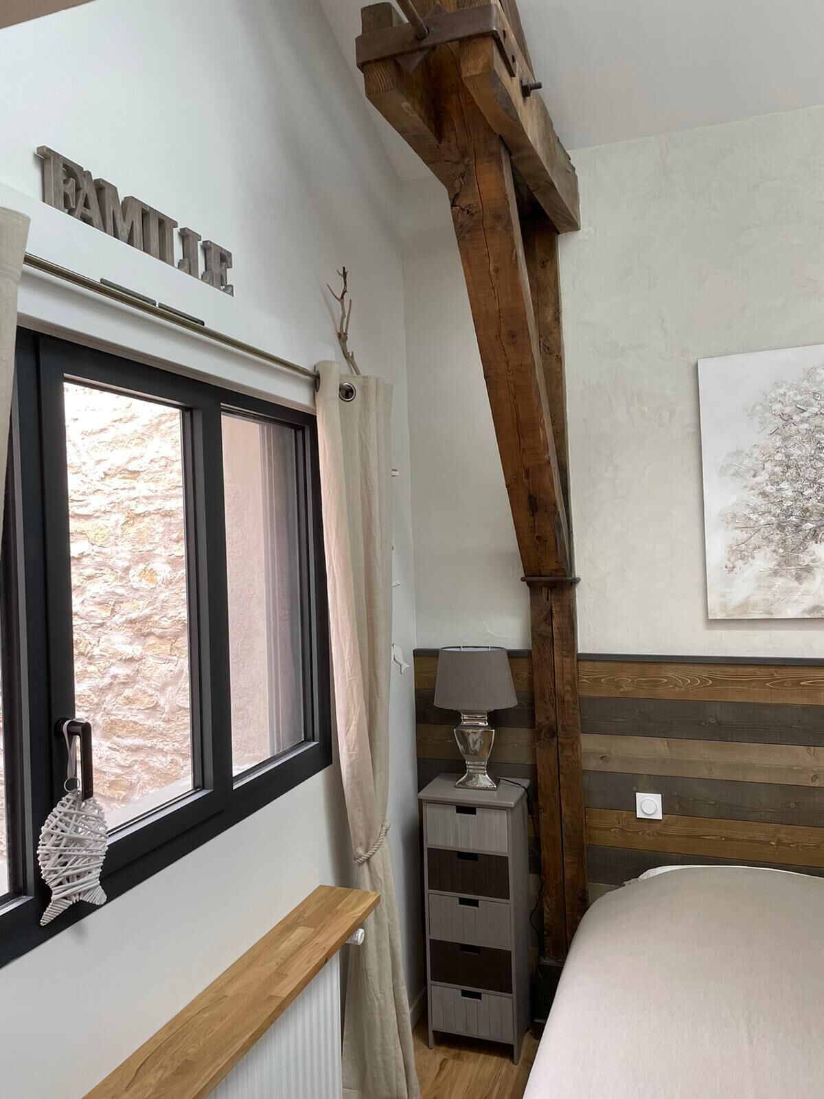 Un dormitorio con ventana, estructura de vigas de madera, una mesita de noche con lámpara y una obra de arte en la pared.